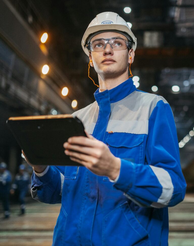 Opportunities Engineer in safety gear uses a tablet in a large industrial factory setting, focused and diligent.